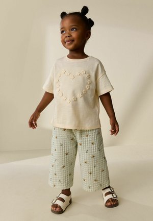 Lille barn med et smil iført en cremefarvet skjorte med et blomsterhjerte og lysegrønne bukser med et honningbi-mønster, stående med sandaler.