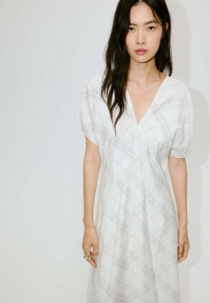 Mujer con cabello largo y oscuro que lleva un vestido blanco con un patrón de cuadros gris claro, de pie frente a un fondo blanco liso.