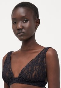 Bralette in pizzo nero con scollatura a V profonda, motivo floreale, spalline larghe e una morbida texture trasparente. Accentuato da un bordo solido.