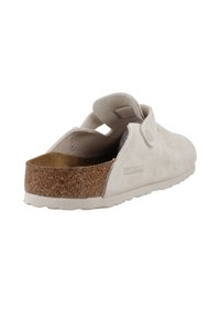 Chaussure slip-on en suède beige avec une semelle intermédiaire en liège, une sangle à l'arrière et une semelle en caoutchouc texturé. Elle présente une forme arrondie et des détails de design minimalistes.