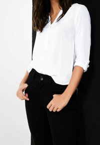 Camicia bianca con colletto henley e maniche arrotolate, abbinata a jeans neri. Tessuto morbido e vestibilità comoda sono evidenti.