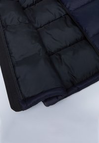 Gros plan sur la manche et le bord avant d'une veste matelassée bleu marine, avec des coutures et une fermeture éclair visibles sur un fond gris clair.