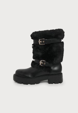 Botte noire mi-mollet avec semelle épaisse en caoutchouc, dessus en fausse fourrure et deux sangles en cuir réglables avec boucles métalliques.