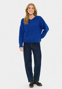 Blauwe gebreide trui met een V-hals, losse pasvorm en ribgebreide manchetten, gecombineerd met donkere rechte jeans en zwarte schoenen. Effen achtergrond.