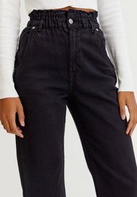 Persoon in hoge zwarte broek met gathered tailleband en voorzakken, gecombineerd met een witte longsleeve top, handen ontspannen aan de zijkanten.