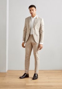 Completo beige chiaro con una vestibilità su misura, tessuto strutturato e due bottoni. Camicia bianca sotto il completo, scarpe eleganti marroni, in piedi su un pavimento di legno.