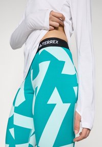 Leggings turquoise avec de grands motifs de flèches blanches, dotés d'une taille noire TERREX. Le matériau semble extensible avec une texture lisse.