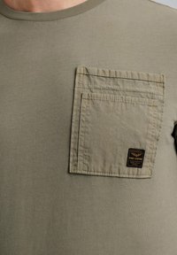 Camiseta de manga corta color verde oliva con un bolsillo en el pecho texturizado y sobredimensionado y una etiqueta. Material de algodón suave con un escote redondeado.