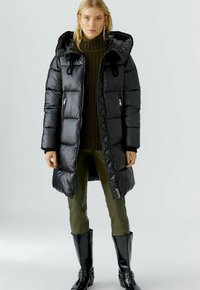 Zwarte pufferjas met een capuchon, gemaakt van glanzend materiaal, met ritszakken en een knielange stijl. Gedragen over een donkergroene trui en broek.