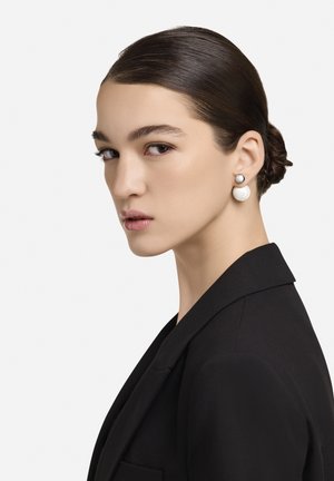 Biserna uhana z bleščečim zaključkom; ena ima bel okrogel obliko, druga pa srebrn kovinski poudarek. Nosi jih modelka s sijočimi lasmi.