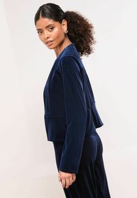 Navy plavi baršunasti skraćeni sako s dugim rukavima, peplum dizajnom i teksturiranim završetkom, u kombinaciji s hlačama od istog materijala.