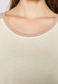 Kortärmad skjorta i krämfärg med rund halsringning som har en kontrasterande mjuk rosa kant. Tyget verkar slätt med en lätt lyster.