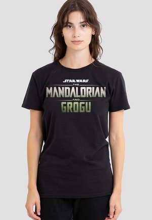 Junge Frau trägt ein schwarzes "Star Wars The Mandalorian and Grogu" T-Shirt und steht vor einem einfarbigen hellgrauen Hintergrund.