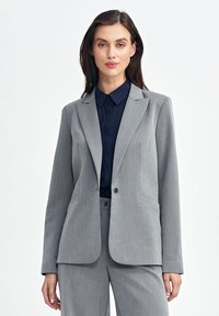 Szary dopasowany żakiet z pojedynczym zapięciem na guzik, ostrymi klapami, bocznymi kieszeniami i gładką teksturą, w połączeniu z ciemnoniebieską koszulą z kołnierzykiem.