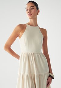 Nainen ilman hihoja beiges mekossa, jossa on kerroksellinen hame, isojen renkaita korvakorujen ja paksujen rannekorujen kanssa, poseeraa yksinkertaista valkoista taustaa vasten.