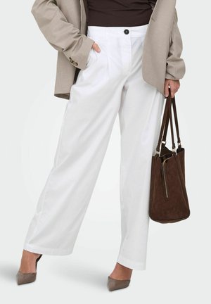 Femme portant un pantalon blanc à jambes larges, un blazer taupe, un haut foncé, des talons taupe, tenant un sac cabas en daim marron foncé à côté d'elle.