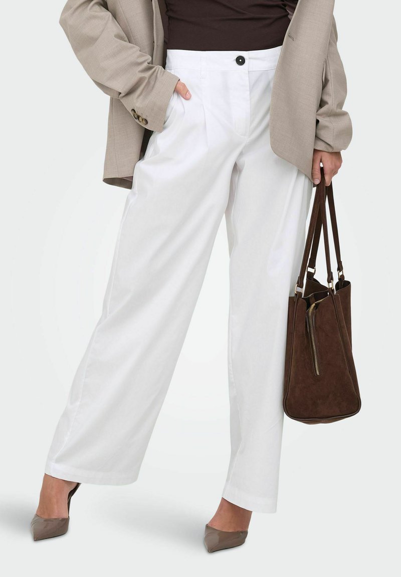 Femme portant un pantalon blanc à jambes larges, un blazer taupe, un haut foncé, des talons taupe, tenant un sac cabas en daim marron foncé à côté d'elle.