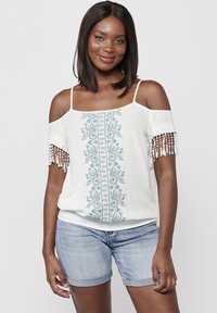 Top bianco off-the-shoulder con dettagli ricamati blu e accenti in pizzo sulle maniche, abbinato a pantaloni denim.
