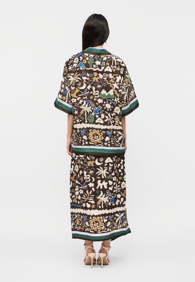 Γυναίκα που φοράει ασορτί φαρδιά μπλούζα και μακριά φούστα με πολύχρωμα αφηρημένα σχήματα, φυτά και ζώα σε σκουρόχρωμο φόντο, όρθια με την πλάτη γυρισμένη.