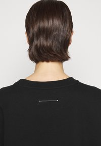 Černý svetr s kulatým výstřihem. Obsahuje malé bílé šití na zadní straně. Hladká textura, minimalistický design.