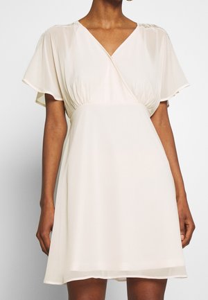 Nainen, jolla on lyhythihainen, V-kaula-aukollinen, vaalean beige kietaisumekko, jossa on hieman levenevä helma ja pitsiyksityiskohdat olkapäillä.