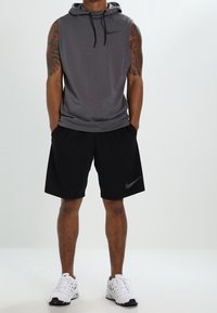 Hoodie gris sans manches avec cordons noirs, associé à un short noir. Logo Nike présent sur le haut et le short. Baskets blanches sportives.