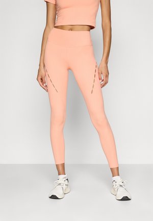 A barack színű leggings sima, rugalmas anyagból készült, magas derékkal és kivágásokkal a külső combokon.
