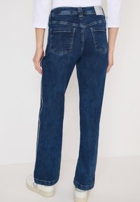 Donkerblauwe wijde jeans met een gladde textuur, zilveren zijkanten en twee achterzakken. Gestyleerd met witte sneakers.