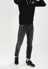 Persona vestida con una sudadera negra, jeans enrollados gris oscuro y zapatillas negras, de pie contra un fondo claro y liso.