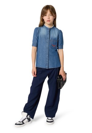 Chemise en denim à manches courtes bouffantes, fermeture à boutons sur le devant, poche avec détails rouges. Associée à un pantalon ample bleu marine et un sac à main noir.