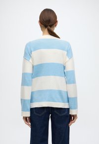 Gaiši zils un krēmkrāsas svītrains džemperis ar ribotu apaļu kaklu un aprocēm, mīksta tekstūra un vaļējs siluets, kombinēts ar tumša džinsa biksēm.