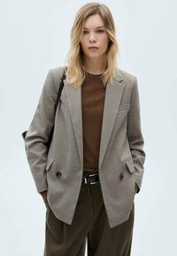 Blazer doppiopetto in un motivo pied de poule, tonalità di grigio e marrone, con rever a punta e grandi bottoni, indossato sopra un maglione marrone.