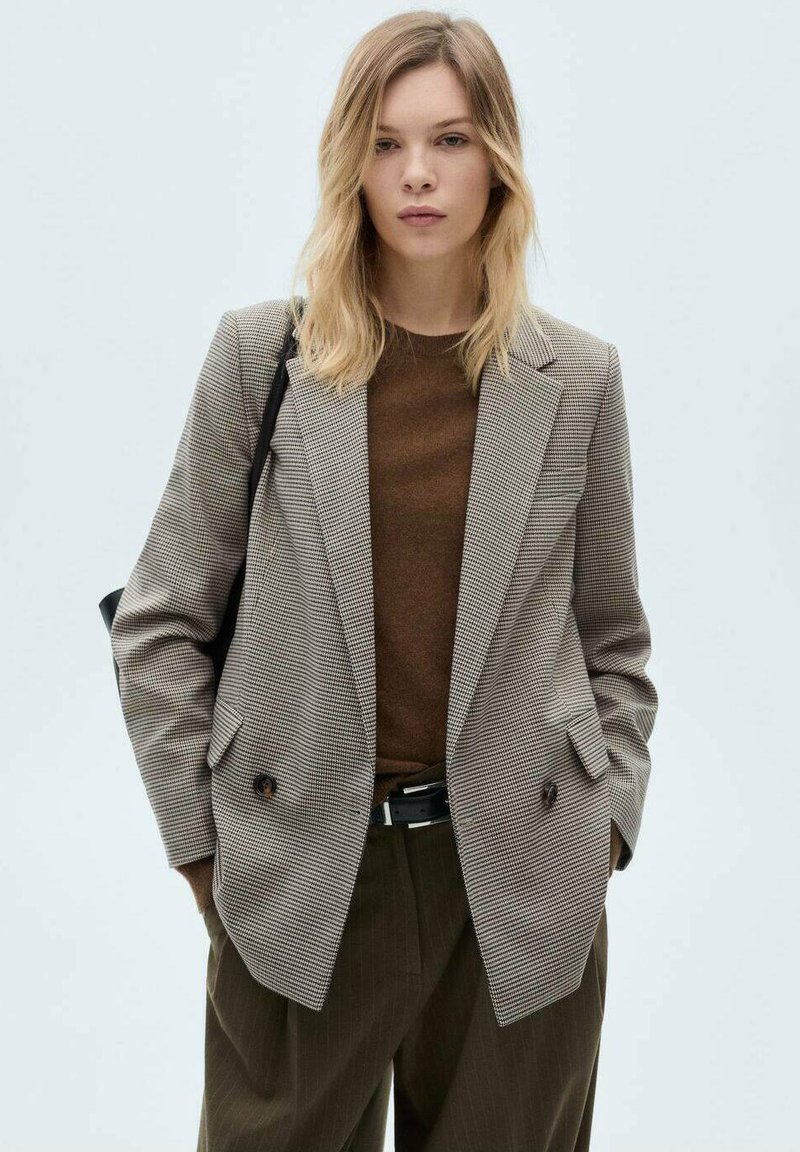 Blazer doppiopetto in un motivo pied de poule, tonalità di grigio e marrone, con rever a punta e grandi bottoni, indossato sopra un maglione marrone.