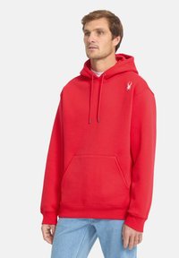 Sweatshirt à capuche rouge en tissu doux, avec une poche kangourou, des cordons de serrage et un petit logo blanc sur la poitrine.