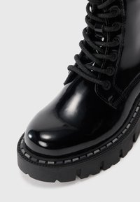 Black patent leather ankle boot with a rounded toe, thick treaded rubber sole, and black laces woven through eyelets, featuring a smooth texture.