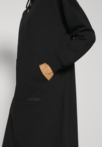 Henkilö, jolla on päällään musta pitkähihainen huppari-mekko, jossa on käsi etutaskussa ja pieni "Marc O'Polo" -logo.