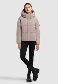 Gepolsterte hellbeige Jacke mit hohem Kragen und Kapuze, ausgestattet mit Reißverschluss, gesteppter Textur und Seitentaschen. Schwarze Leggings und Stiefel.