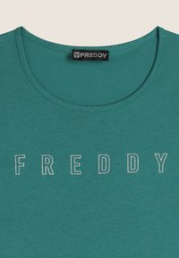 Maglietta a maniche corte di colore teal realizzata in tessuto morbido. Presenta un collo tondo e la scritta argentata "FREDDY" sul petto con un carattere in grassetto.