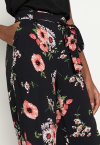 Pantalon noir à motifs floraux avec de grandes fleurs roses et blanches. Ceinture à nouer à la taille et poches sur les côtés. Tissu léger avec une texture lisse.