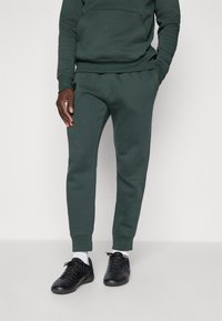 Mørkegrønne joggebukser med slim fit, laget av mykt stoff, med elastisk midje og ankelmansjetter, sammen med svarte treningssko.