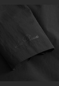 Svart tekstil med en glatt tekstur, med en subtil brodert logo "Les Deux" ved mansjetten i en matchende svart tråd.
