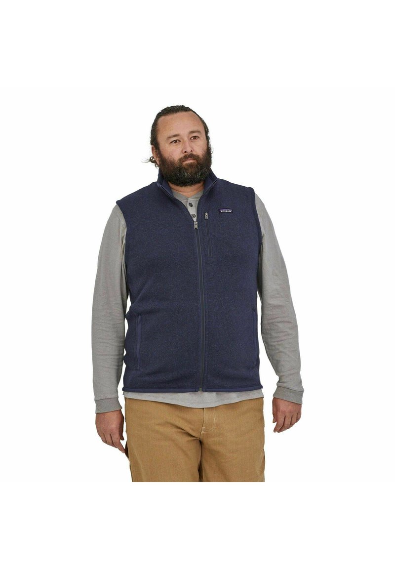 Chaleco de forro polar azul marino con cierre de cremallera, cuello alto y dos bolsillos laterales. Llevado sobre una camisa de manga larga ligera y combinado con pantalones caqui.