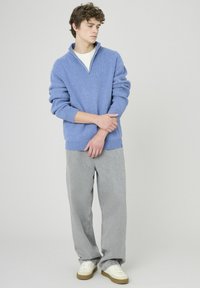 Jeune homme portant un pull bleu à fermeture éclair quart, une chemise blanche, un pantalon gris ample et des baskets blanches, se tenant devant un fond uni.