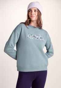 Light blue sweatshirt with ribbed cuffs and hem, featuring "Après" in a floral print. Paired with dark purple leggings and a lilac beanie.