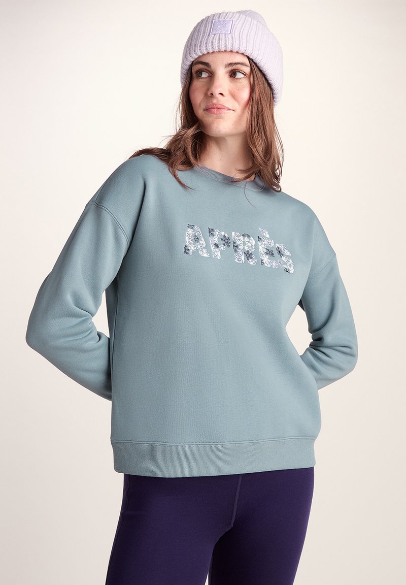 Light blue sweatshirt with ribbed cuffs and hem, featuring "Après" in a floral print. Paired with dark purple leggings and a lilac beanie.