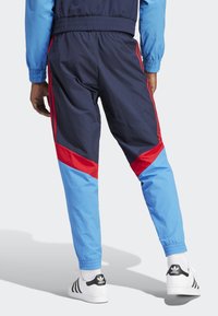 Marineblaue Windjackenhose mit roten Akzenten, elastischen Bündchen und schmalem Schnitt. Kombiniert mit weißen Turnschuhen mit schwarzen Streifen.