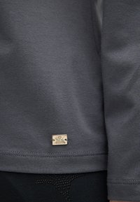 Šedá mikina s hladkým povrchem, se zlatou logem na lemu. Švy jsou viditelné podél okrajů.