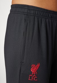 Juodos sportinės kelnės su elastinga juosmena, turinčios šonines kišenes ir raudoną siuvinėtą "Liverpool FC" logotipą kairiajame šlaunyje.