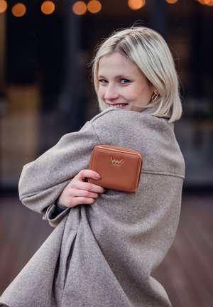 Brown leather wallet with a zip closure, featuring a subtle embossed logo, held against a light gray, textured coat.