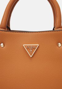 Sac à main tan en cuir texturé, avec des accents en métal doré et un badge logo triangulaire portant "GUESS USA" à l'avant.